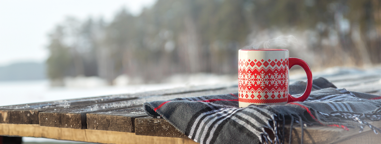 A cup with a festive design behind a snow landscape background with trees.