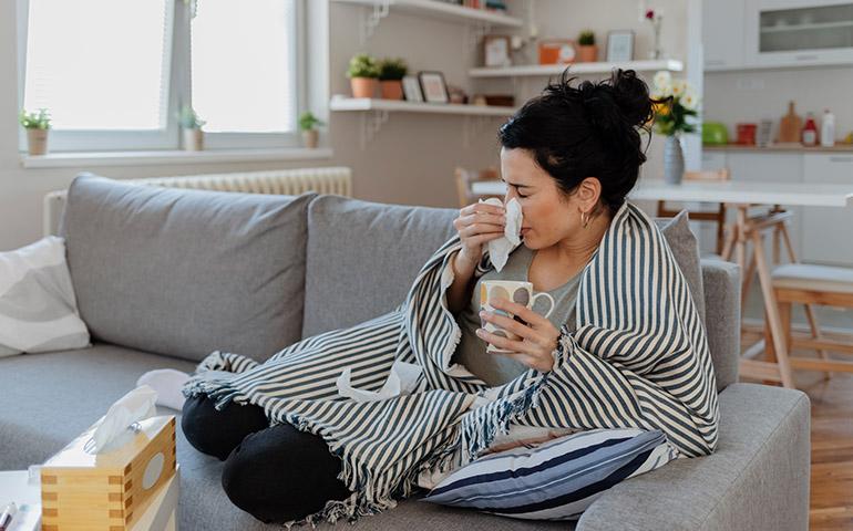 Woman sitting on a couch wrapped in a blanket sneezing into a tissue